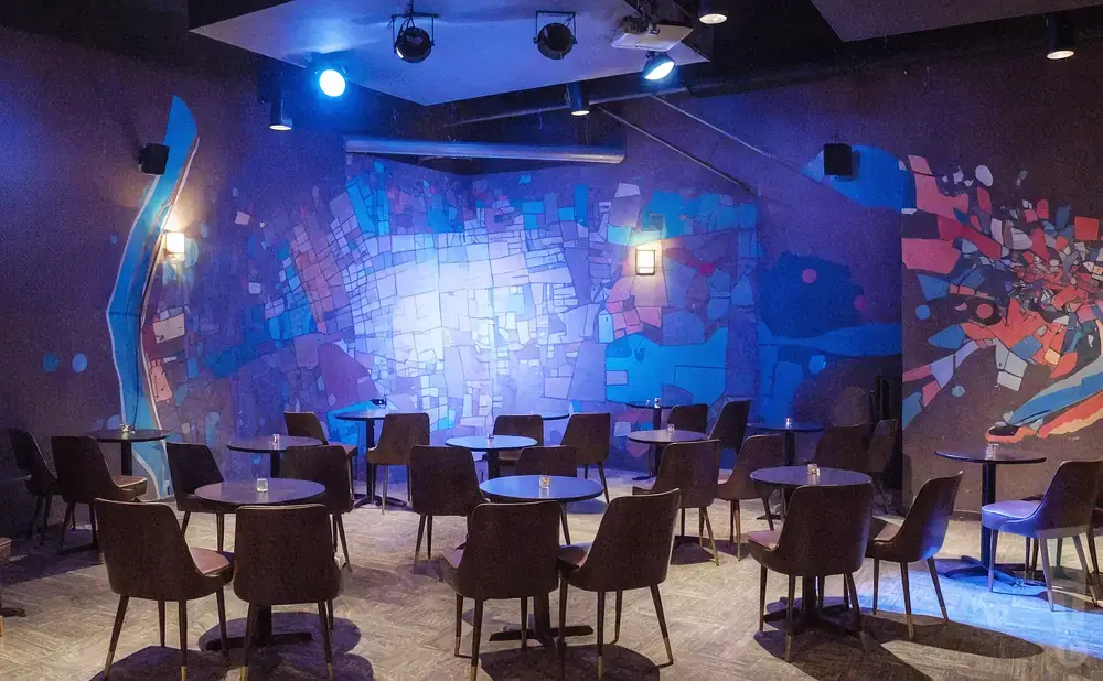 Dark room with colorful abstract murals, tables, and chairs, lit by blue spotlights.