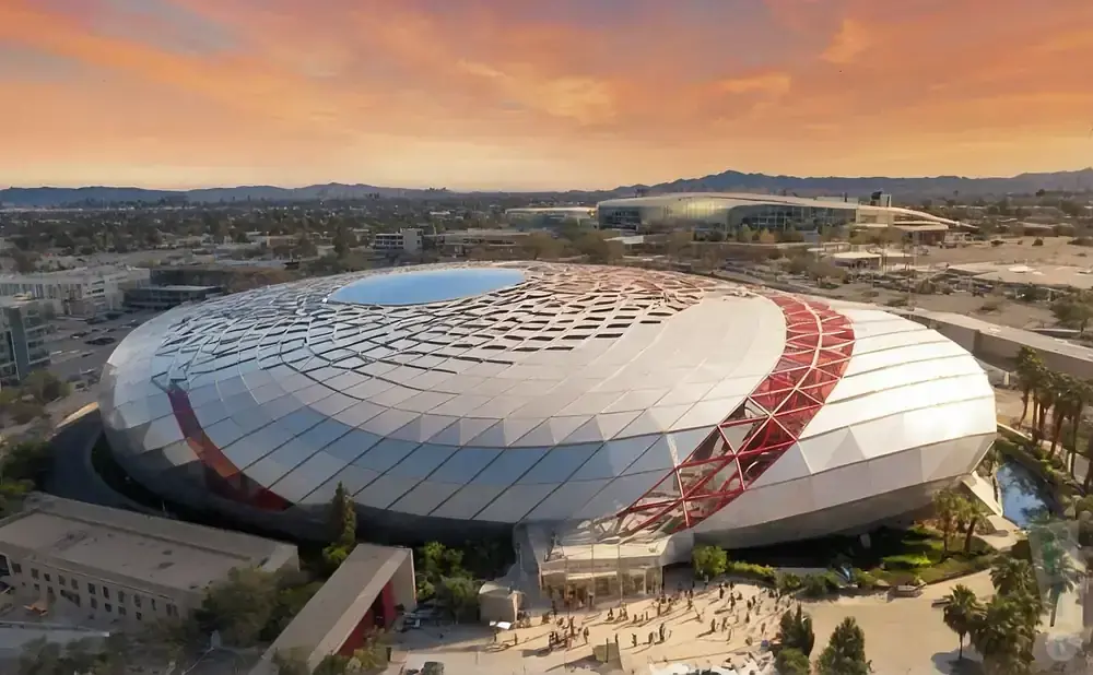 Aerial view of a modern, circular stadium with a patterned, white roof and red accents, set against a sunset sky.