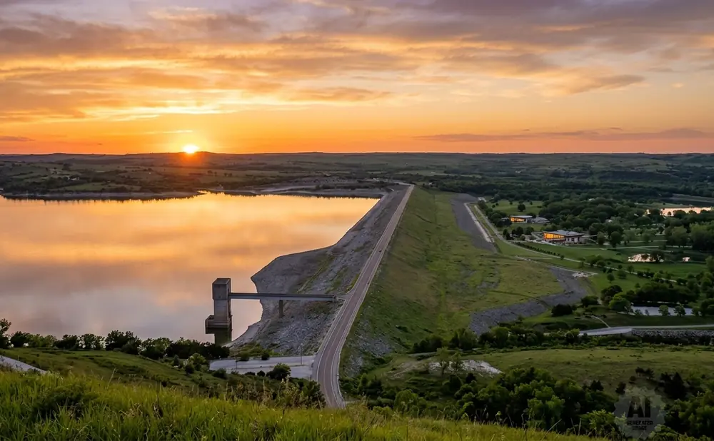 Sunset over a lake and dam, with rolling green hills and trees.