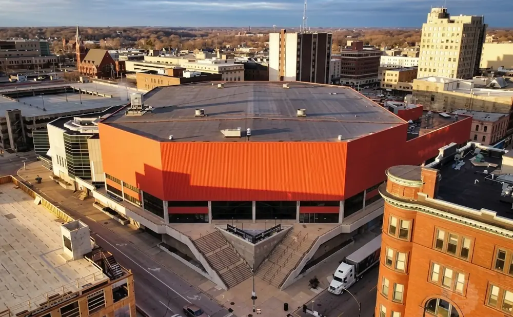 An aerial view of a modern, orange-walled building with tiered concrete stairs leading to its entrance, set against a city skyline.