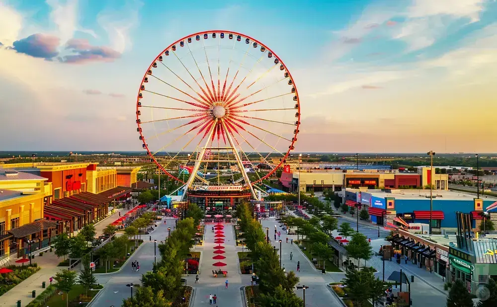 An aerial view of a large Ferris wheel at a lively outdoor entertainment district with shops and restaurants.