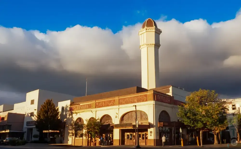 A pale tower with a domed top rises above a tan building with arched windows, under a blue sky with white and dark clouds.