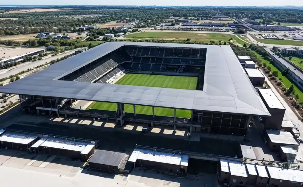 Aerial view of a modern, rectangular stadium with a large gray roof and a vibrant green soccer field.