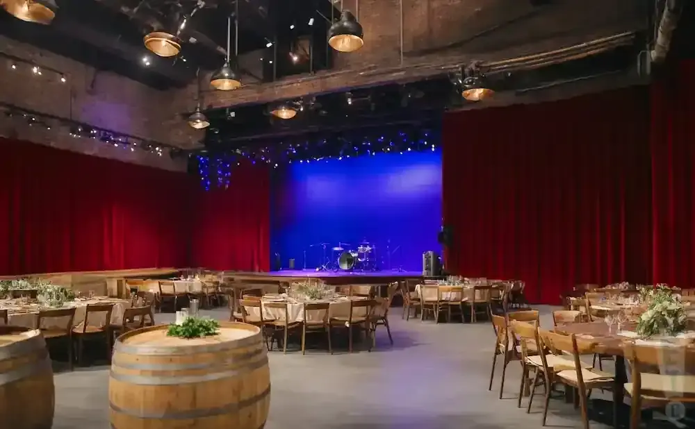 A venue set up for an event with tables and chairs, a stage with a blue backdrop, and red curtains.