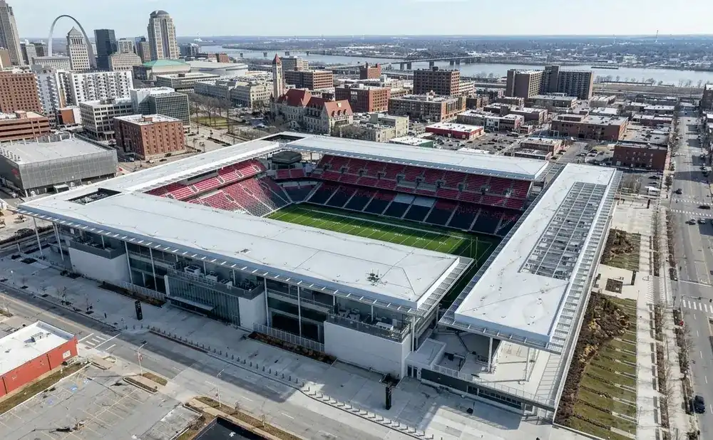 Aerial view of a modern stadium with red and black seats, a green field, and surrounding city buildings.
