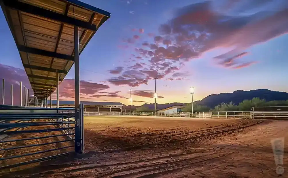 Equestrian arena at sunset with dramatic clouds, lights, and mountains in the background.