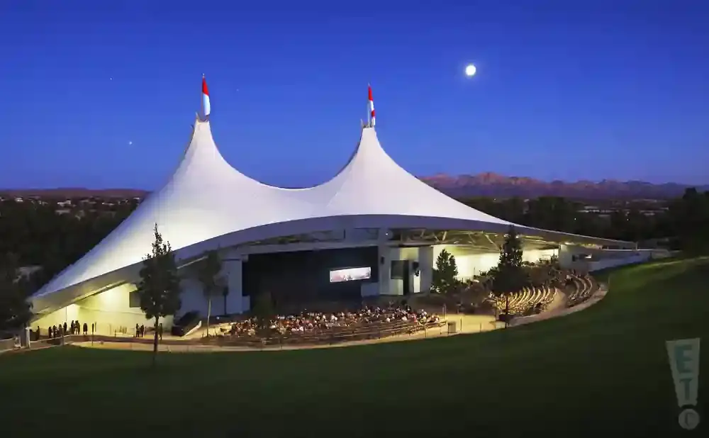 An outdoor amphitheater with a white tent roof at dusk, with mountains in the distance and a full moon in the sky.