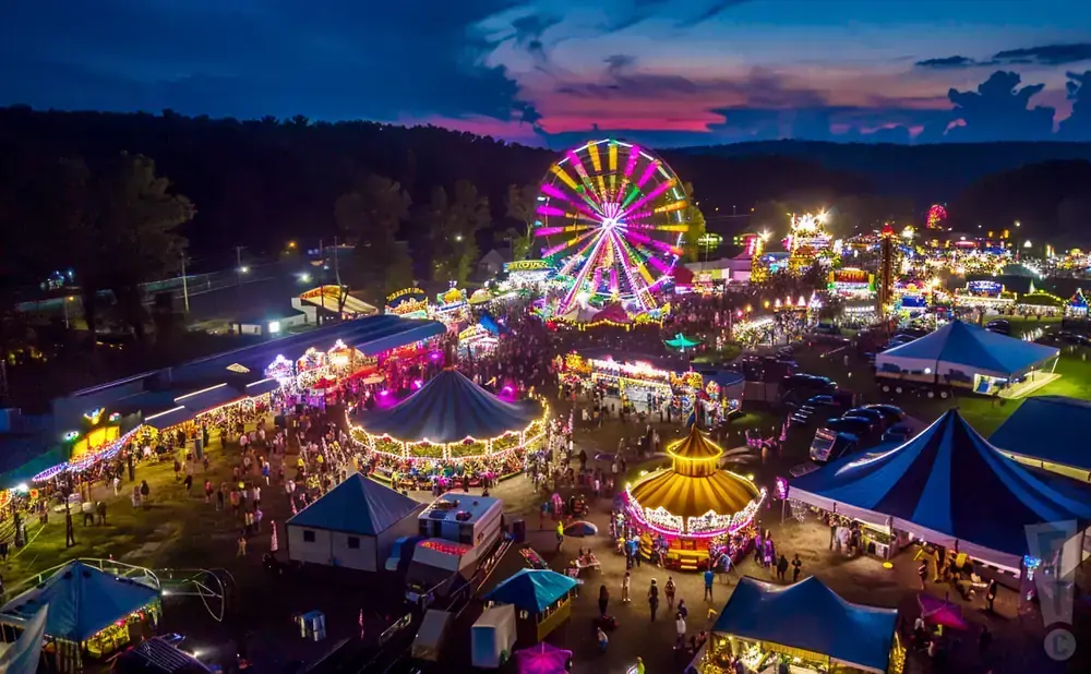 An aerial view of a brightly lit carnival at dusk with a Ferris wheel, carousels, and crowds of people.
