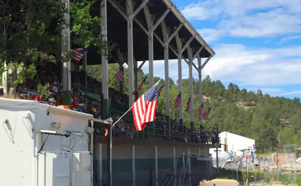 American flags wave in front of bleachers filled with spectators at an outdoor event.