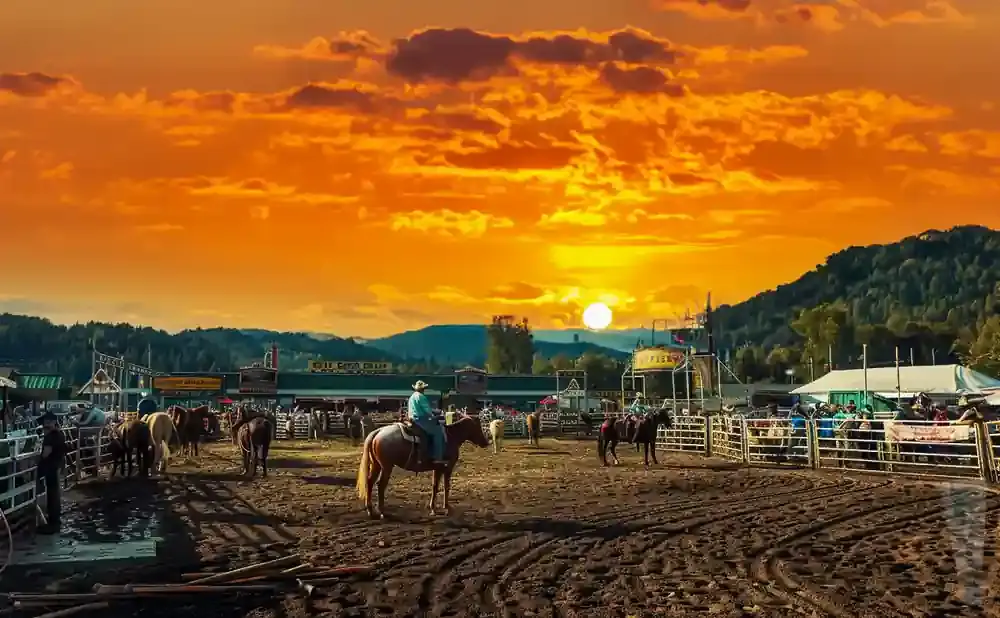Rodeo arena at sunset with cowboys on horses and a vibrant orange sky.