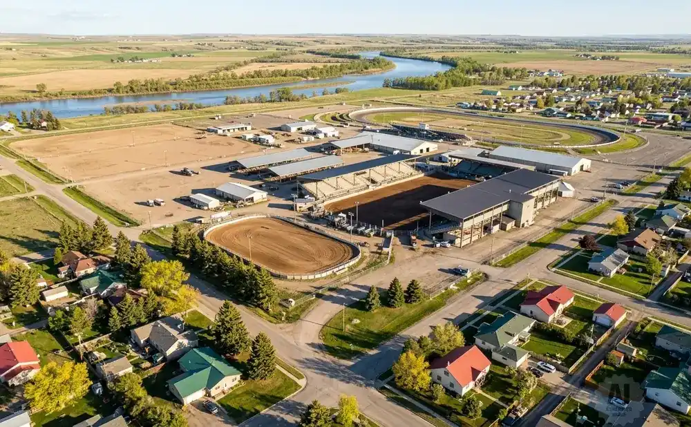 Aerial view of a rodeo arena and grandstands next to a curving racetrack, with a river and rolling hills in the background.