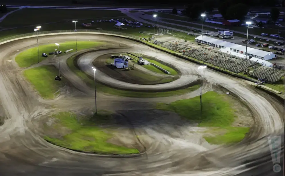 Aerial view of a dirt race track at night, with lights illuminating the track and grandstands.