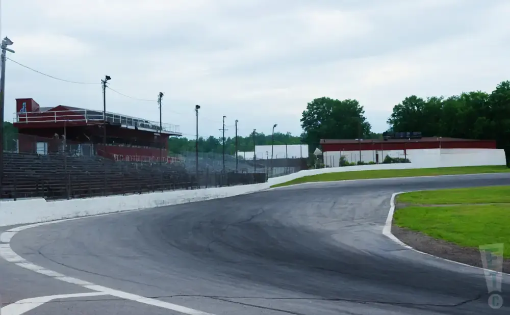 A race track with a grandstand, buildings, and trees under a cloudy sky.
