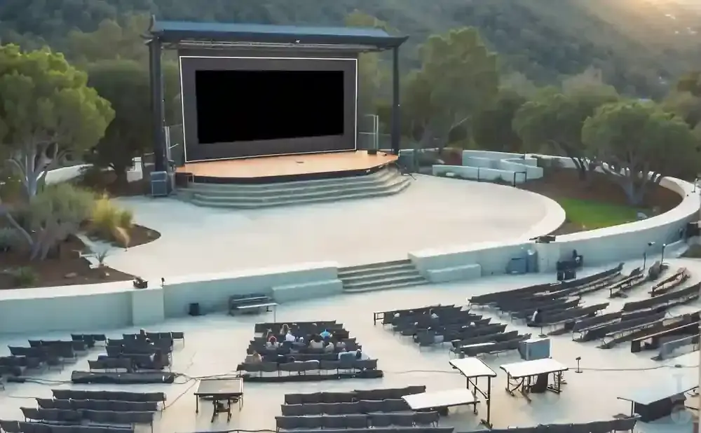 An outdoor amphitheater with a stage and screen, facing rows of empty seats and a few scattered audience members.