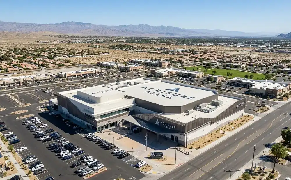 Aerial view of Acrisure Arena in Palm Springs, CA, surrounded by parking lots and commercial buildings, with mountains in the background.