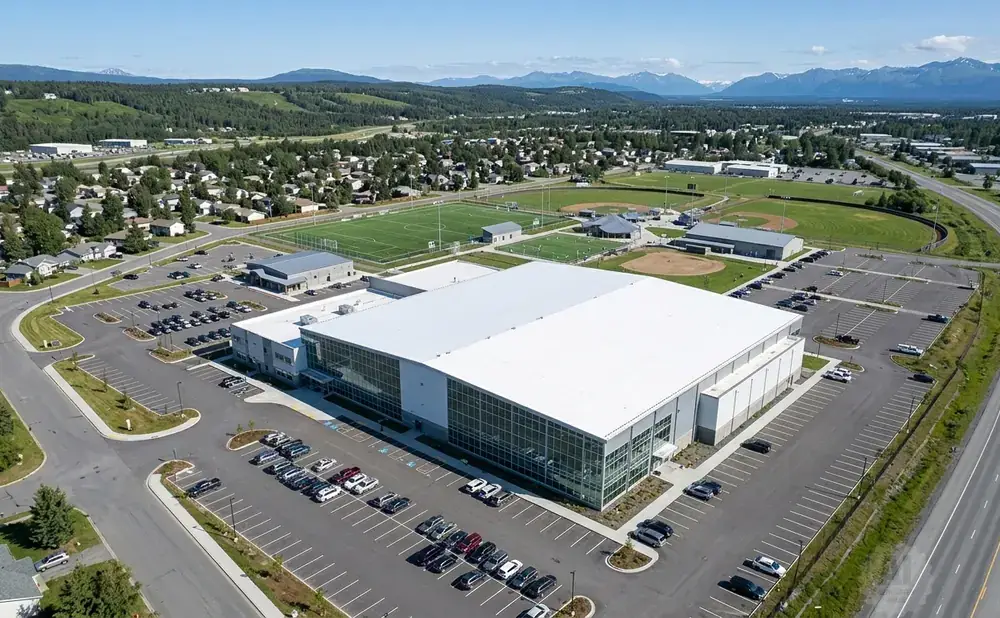 Aerial view of a modern indoor sports complex with large windows, surrounded by parking lots, sports fields, and residential areas.