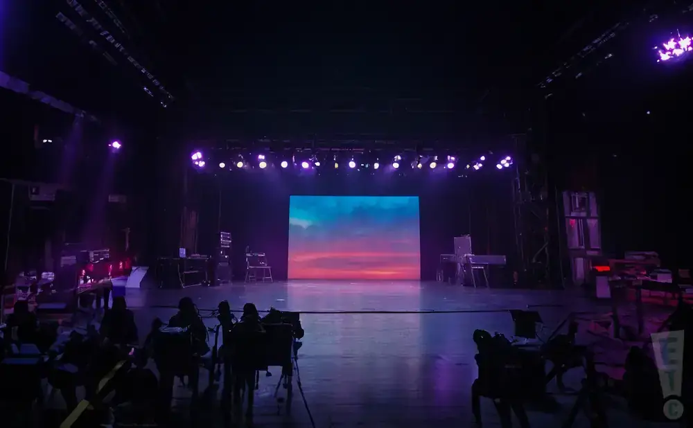A stage with a large screen displaying a sunset, purple lights, and silhouettes of people in the foreground.
