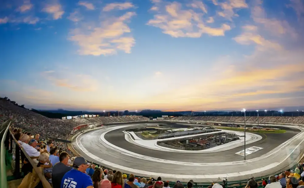 Spectators watch a race at a speedway under a sunset sky with clouds.