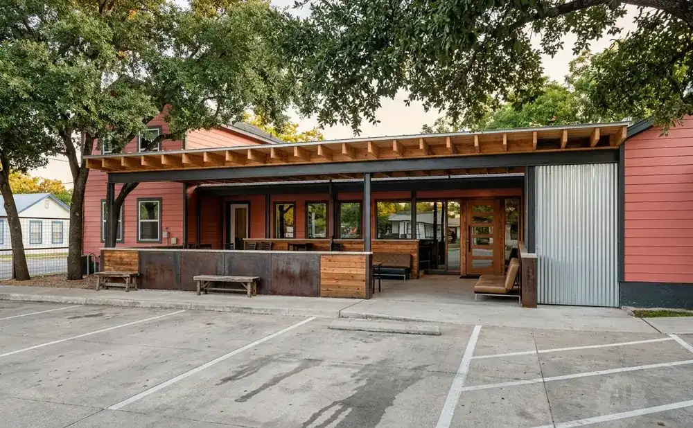 Exterior view of a salmon-colored building with a covered outdoor patio and seating area, facing a parking lot.