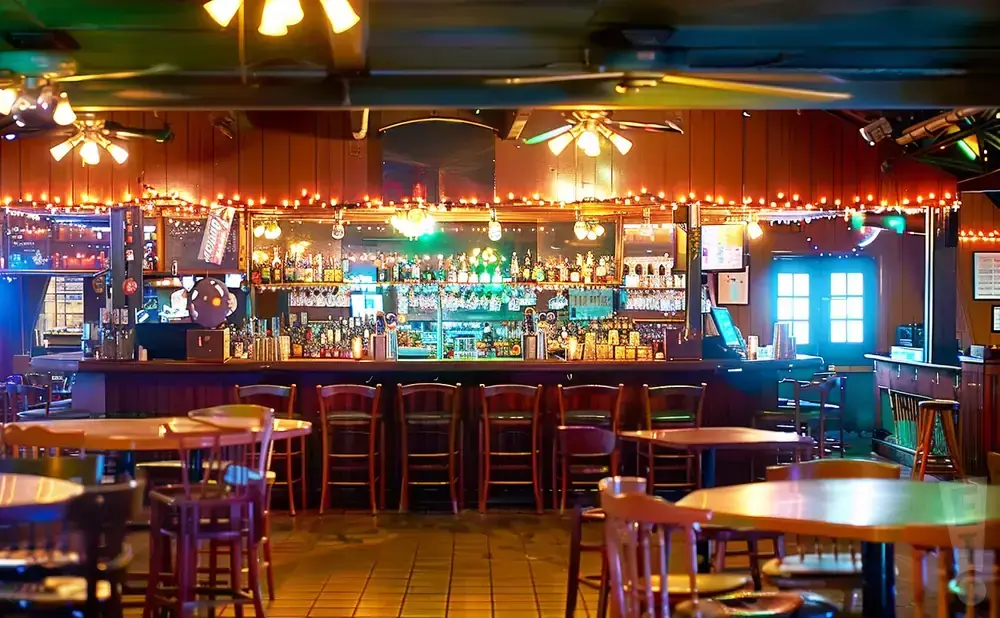Interior of a dimly lit bar with wooden stools, tables, and a well-stocked liquor shelf illuminated by string lights.