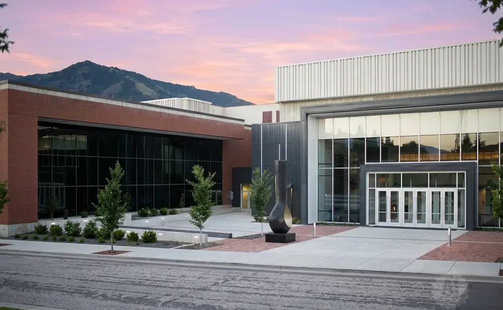 Modern building with brick and glass facade, large windows reflecting mountains under a pink sky.