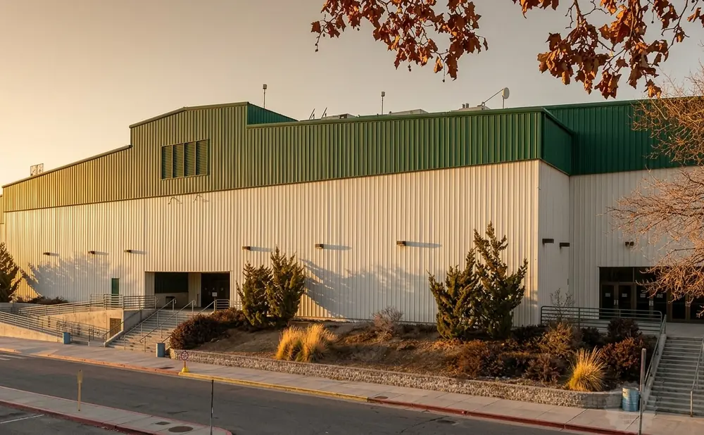 A large white and green building with sloped roofs, illuminated by warm evening light, with trees and a street in the foreground.