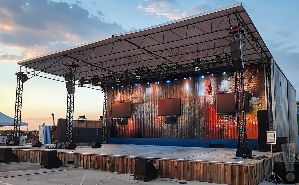 Outdoor stage setup with a corrugated metal backdrop, screens, speakers, and lighting.