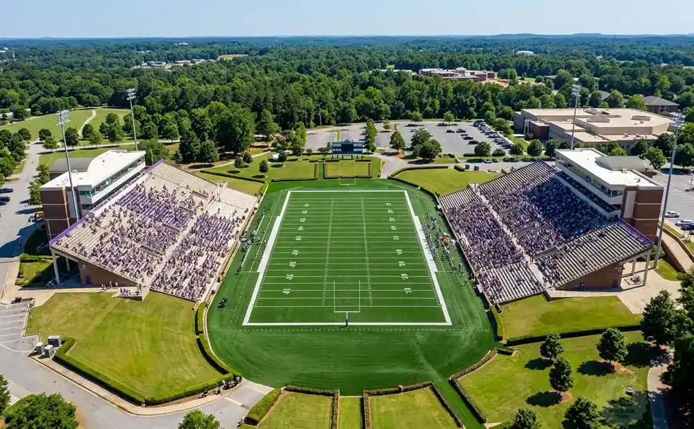 Aerial view of a football stadium with purple-seated stands full of fans, a green field with white yard lines, and surrounding trees and buildings.