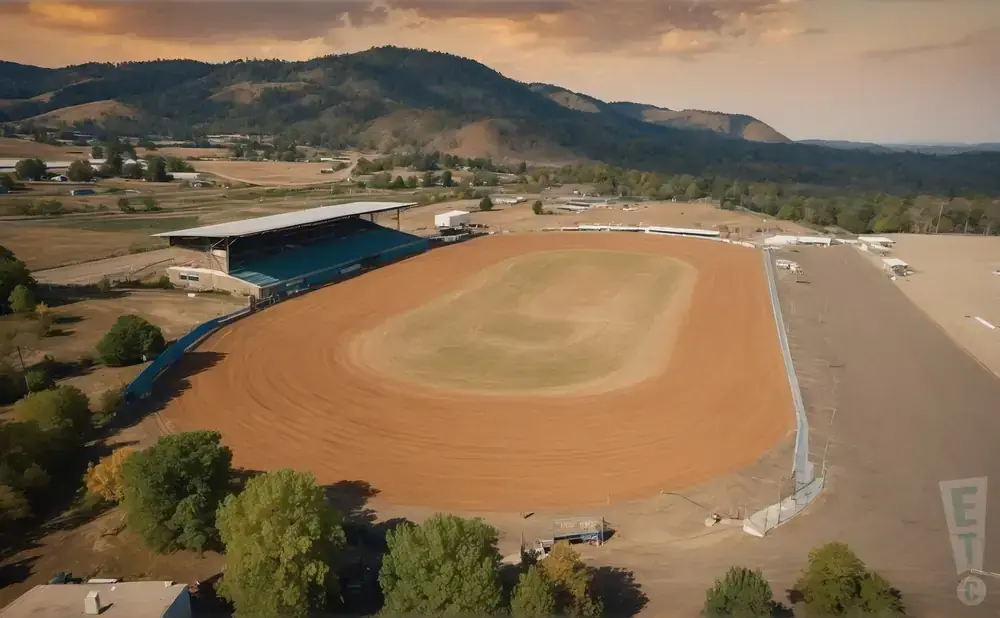 Aerial view of a dirt oval racetrack with grandstands and surrounding hills.