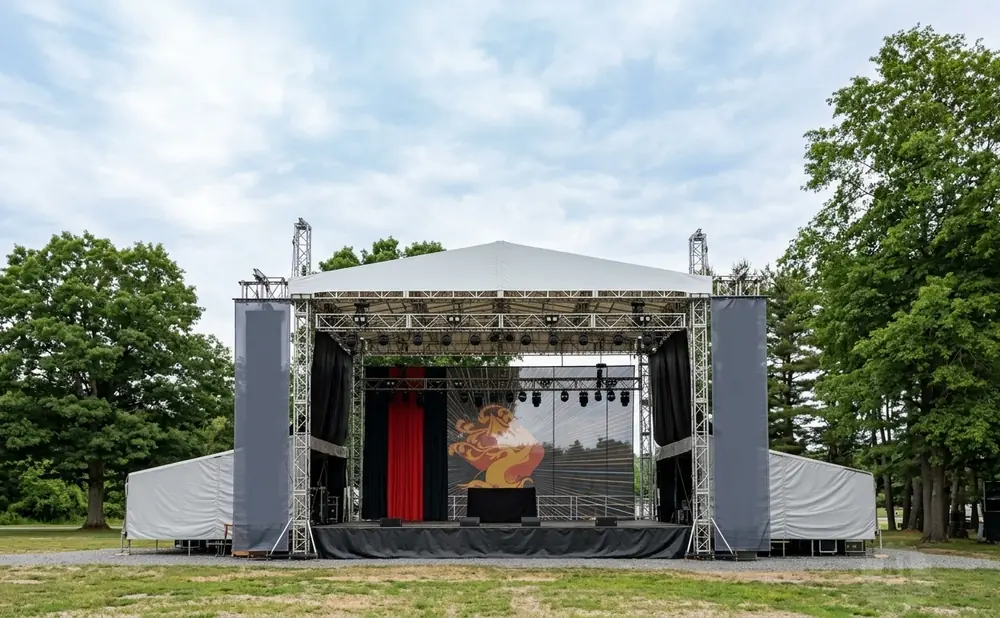 An outdoor stage setup with a white tented roof, trussing, speakers, and a large screen displaying a graphic.