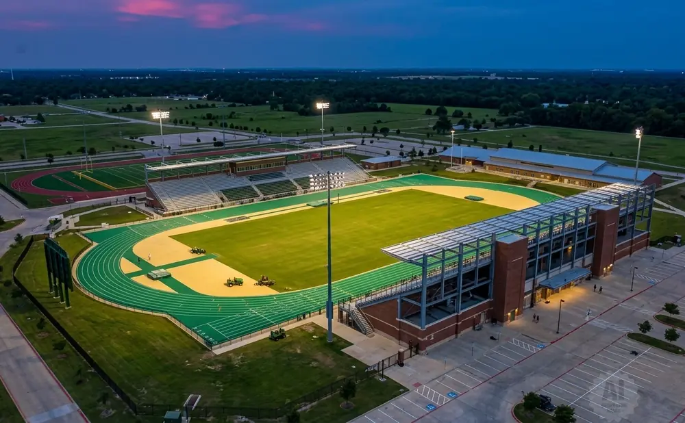 Aerial view of a modern sports stadium complex at dusk, featuring a football field, track, and bleachers.