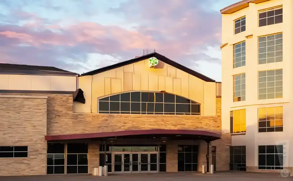A modern building with stone and light-colored facade, featuring large windows and a prominent green logo on the roof against a sunset sky.