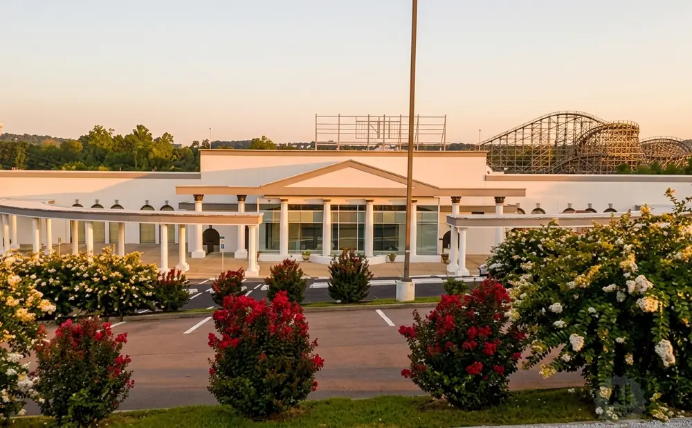 Exterior of a white building with a covered walkway, surrounded by blooming bushes and a roller coaster in the background.