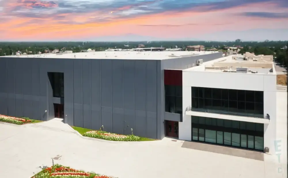 Modern industrial building with gray walls and white accents, featuring large windows and colorful flower beds.