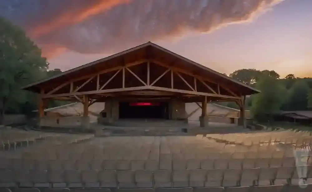 Outdoor amphitheater with rows of empty seats facing a wooden stage, under a dramatic sunset sky.