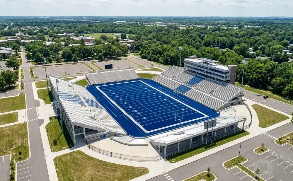 Aerial view of a blue football field with stadium seating surrounded by trees and buildings under a cloudy sky.