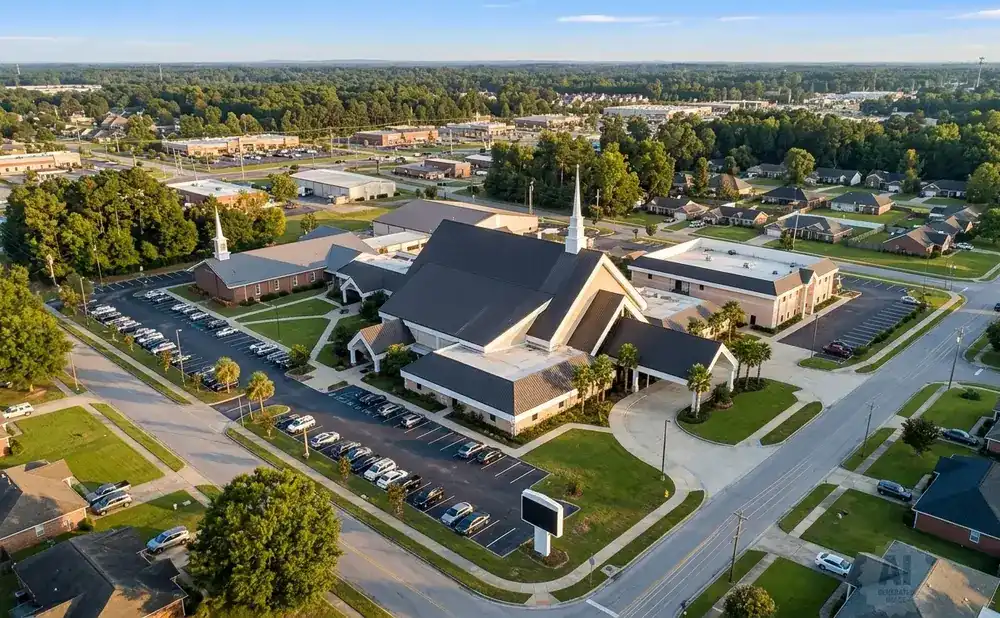 Aerial view of a church and parking lot, surrounded by trees and residential buildings.