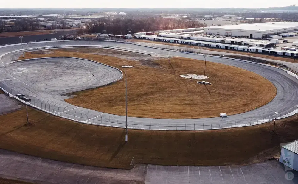Aerial view of a race track with brown grass and a large industrial building in the background.