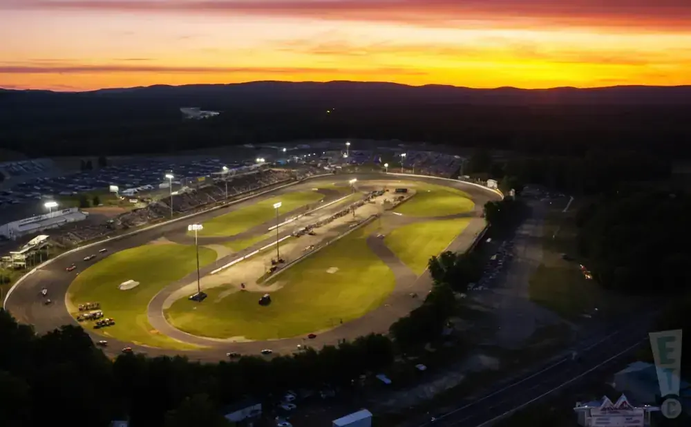 Aerial view of a race track at sunset with a vibrant orange and pink sky, floodlights illuminating the track.