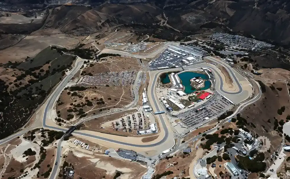 Aerial view of a winding race track with a turquoise lake and grandstands.