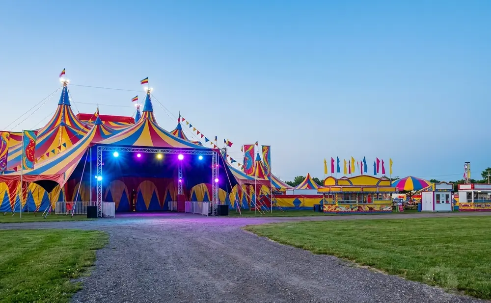 A large, colorful circus tent with a stage, surrounded by smaller tents and food stands at a fair.