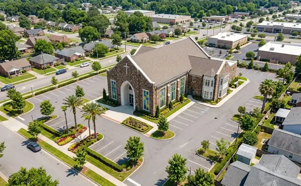 Aerial view of a stone church with colorful stained-glass windows and a parking lot, surrounded by residential homes and businesses.