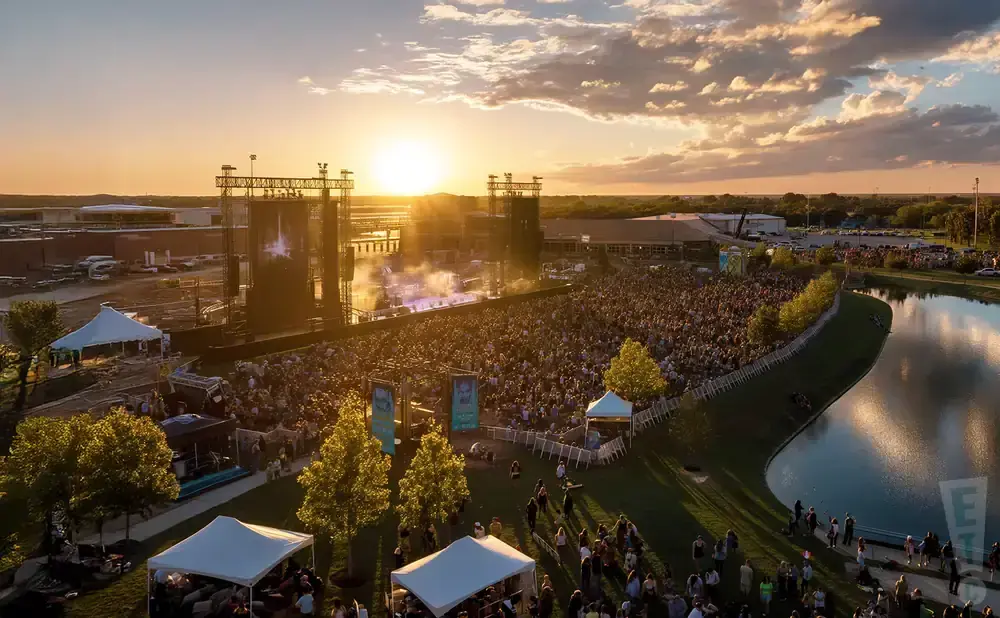 An aerial view of a crowded outdoor concert at sunset, with a stage, audience, and a reflective lake.
