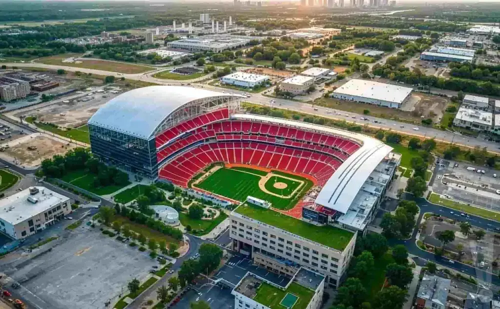 Aerial view of a baseball stadium with red seats and a green field, surrounded by buildings and trees.