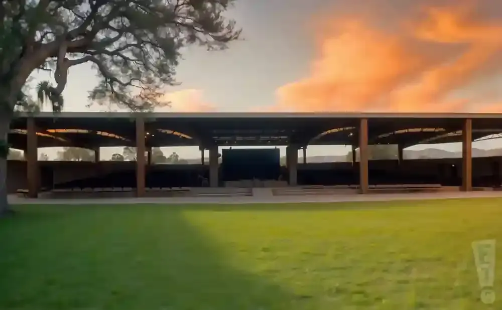 Outdoor amphitheater with seating under a covered pavilion, with dramatic sunset clouds in the sky.