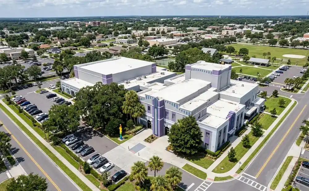 Aerial view of a modern building complex with purple accents, surrounded by trees and parking lots.