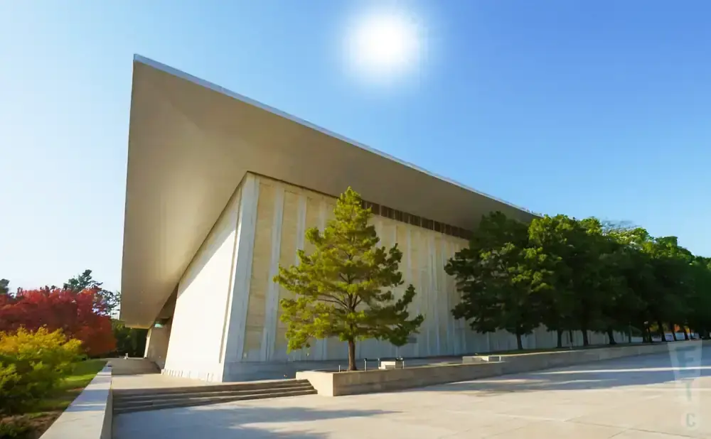 Modern building with a large, angular roof, cream-colored facade, and surrounding trees under a sunny blue sky.
