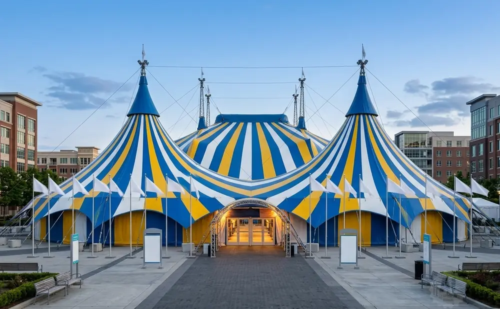 A blue and yellow striped circus tent with white flags in front of modern buildings.