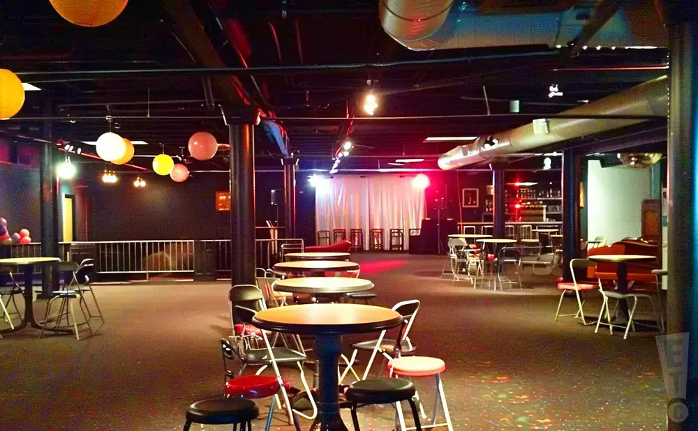 A dark, empty event space with round tables and chairs, lit by colorful lanterns and stage lighting.