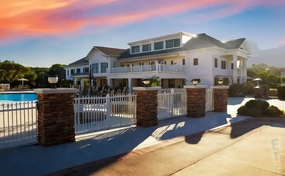 A large white building with columns and a balcony sits behind a white fence with stone pillars.